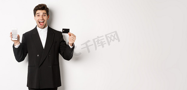 公司形象展示摄影照片_穿着黑色西装的英俊商人的形象，看起来很兴奋，用手机屏幕展示信用卡，站在白色背景下