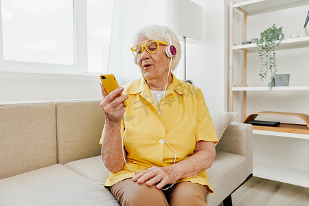 快乐的老妇人带着耳机微笑看着她的电话视频通话、通信技术、明亮的现代室内装饰、生活方式在线通信