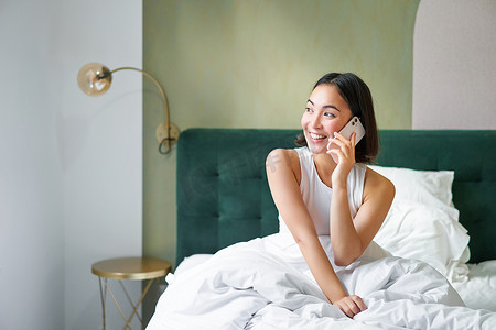美丽的微笑亚洲女孩在手机上聊天，躺在床上，脸上带着高兴的笑脸，在电话里和别人说话