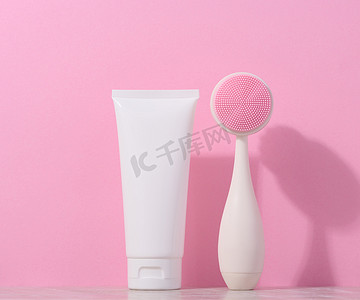 美容霜摄影照片_白色塑料管和粉红色背景中用于面部清洁的按摩刷，用于美容程序的物品