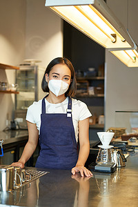 咖啡厅女孩摄影照片_友善咖啡师的垂直照片，戴医用口罩的女职员，在柜台后与客户一起工作，在咖啡厅供应咖啡，穿着制服围裙