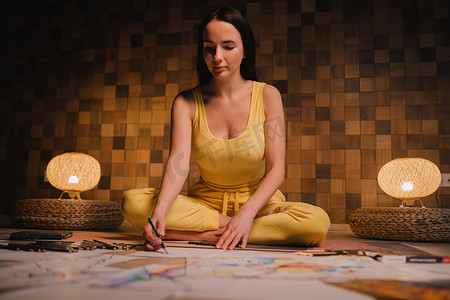 一个穿着黄色衣服的女孩坐在莲花位置上，在家里用晚上的灯光在纸上画画