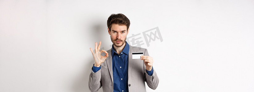 打仗和好摄影照片_穿着西装的快乐男性银行客户展示塑料信用卡和好的标志，微笑着满意，站在白色背景上