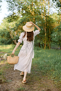 一位身穿夏装长裙的女士在林间小径上行走，手上戴着一顶柳条帽