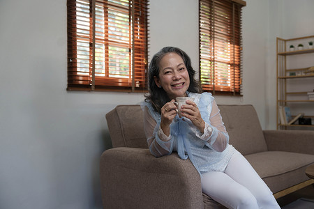 肖像，放松而平静的 60 多岁亚洲女性喝着热茶，在她的客厅里喝下午茶。
