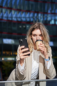 公司照片摄影照片_年轻企业女性的垂直照片喝着咖啡，看着手机、短信，站在城市外面