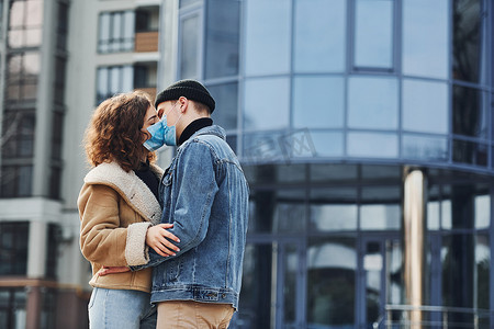 一对戴着防护面具的夫妇在检疫时间在商业大楼附近的城市里互相亲吻。
