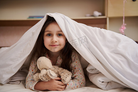 穿着睡衣的可爱小女孩，抱着她的毛绒玩具羊，躺在舒适柔软的白色床上