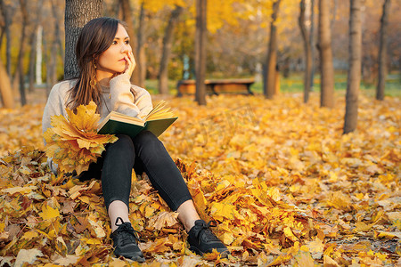 一个年轻女人手里拿着一本书坐在树旁的秋叶上，梦想着什么