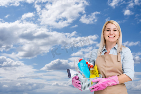 家庭清洁工具摄影照片_年轻女子拿着清洁工具和产品放在桶里，天空背景