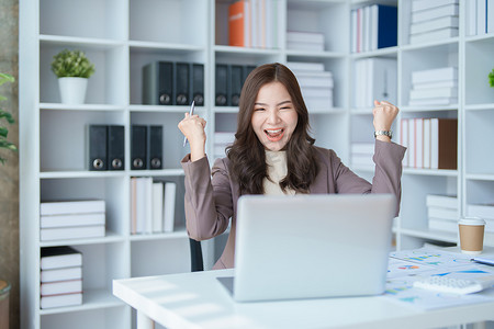 漂亮的年轻亚洲女商人用电脑笔记本电脑举起手来赢取胜利是一种姿态，很高兴成功庆祝成就成功