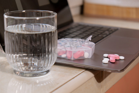 每周摄影照片_组织者每周在工作场所背景和笔记本电脑上用一杯水拍摄医疗药盒的特写镜头，里面装有药片，每天服用白色粉红色药物和胶囊。