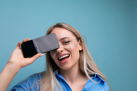 一个快乐的欧洲金发女孩的特写镜头，她拿着智能手机屏幕，在蓝色背景下，她的脸附近有一个应用程序模型