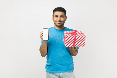 开朗迷人的胡子拉碴的男人，带着礼物盒，展示着空空如也的手机。