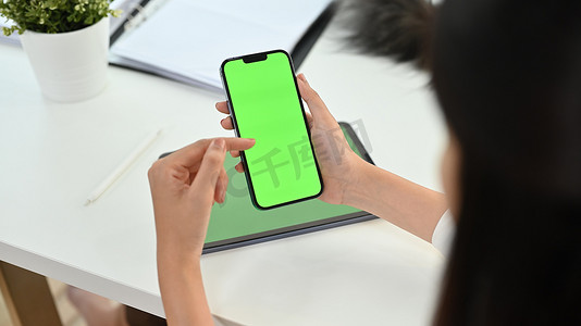 手机办公办公摄影照片_裁剪拍摄的女人拿着智能手机与办公桌上的绿色铬屏幕。