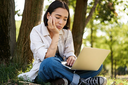 在阳光明媚的夏日，亚洲女性坐在树边的草地上，使用笔记本电脑，工作，远程做作业的肖像