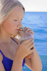 欧洲杯券摄影照片_一个留着欧洲外表的金发的年轻女孩，在海滩的背景下，一个少年手里拿着并喝着一杯彩色的冷无酒精鸡尾酒