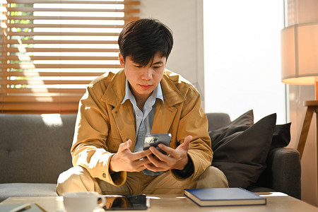 帅气的亚洲男人坐在家里的沙发上，在智能手机的应用程序中在线交流