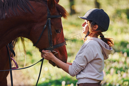 阳光明媚的白天，戴着防护帽的年轻女子和她的马在森林里
