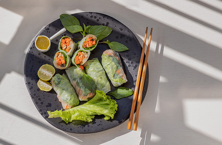 越南菜摄影照片_健康的越南春卷作为蔬菜的一部分，配上辣酱和黑色陶瓷盘上的木筷子。