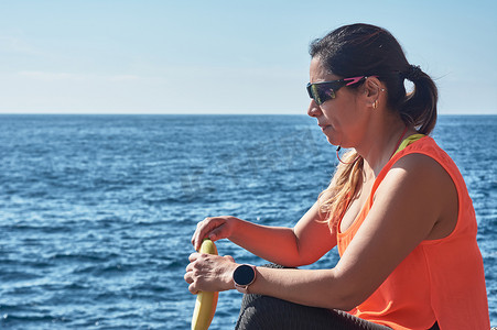 拉丁女人，中年，休息，恢复体力，吃饭，喝水，健身后，燃烧卡路里，保持健康，在海边户外，戴着耳机和智能手表