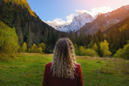 后视，一位身穿红色外套的金发女子站在雪山和茂密的绿色森林的背景下