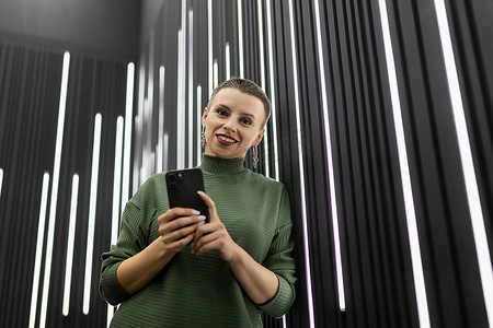 led灯带摄影照片_带垂直 LED 灯的黑墙附近有手机的女人