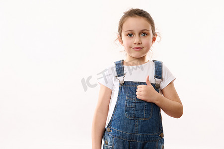 儿童竖起大拇指摄影照片_穿着蓝色牛仔服的漂亮小孩，竖起大拇指，微笑着看着镜头，在白色背景中被隔离