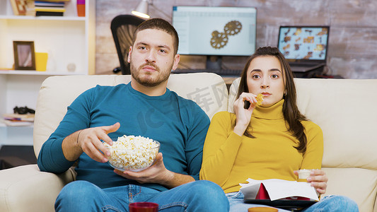 炸鸡实景摄影照片_惊恐的夫妇坐在沙发上看电视吃披萨和爆米花