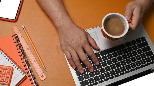男人拿着咖啡摄影照片_男人拿着咖啡杯，在棕色皮革上使用笔记本电脑。