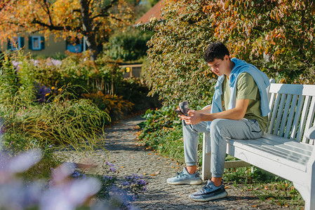 公园里的长椅摄影照片_一个少年坐在秋天公园的长椅上，喝着保温杯里的咖啡，看着手机。
