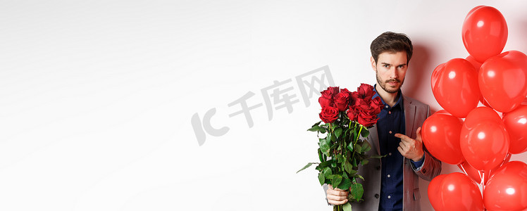 穿着西装的帅哥在浪漫的情人节约会，指着一束玫瑰，站在白色背景上的心形气球旁