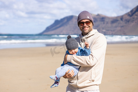 在西班牙兰萨罗特岛法马拉多风的沙滩上，父亲享受着纯净的大自然，抱着他的婴儿男婴晒太阳。