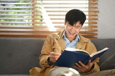 闲暇时光摄影照片_愉快的亚洲男人在沙发上看书，在舒适的周末在家里度过闲暇时光。