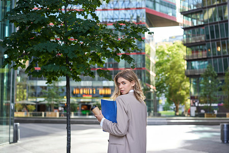 穿着米色西装的年轻女商人的剪影，走在市中心，拿着工作文件和文件，站在办公楼附近的户外
