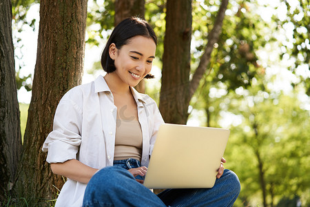 快乐的亚洲小女孩坐在树边的公园里，看着笔记本电脑，在户外远程工作，与人交谈，视频聊天