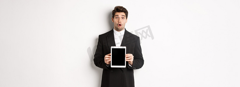 身穿黑色西装的帅哥惊讶的形象，展示数字平板电脑屏幕，看起来很惊讶，站在白色背景下