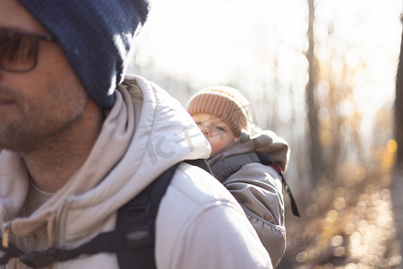 冬季大花连衣裙子摄影照片_运动型父亲背着他的小儿子穿着冬季连身衣和帽子，背着背包在秋天的森林里徒步旅行。