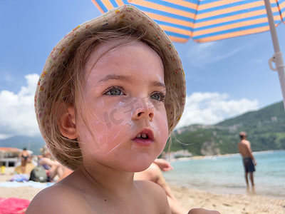 脸上涂防晒霜的小女孩坐在太阳伞下的沙滩上