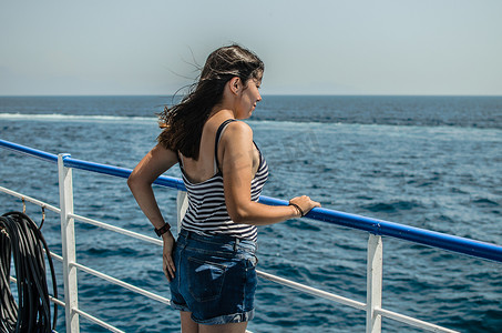 牛仔美女摄影照片_穿着条纹T恤和蓝色牛仔短裤的黑发美女站在海上一艘船的甲板上