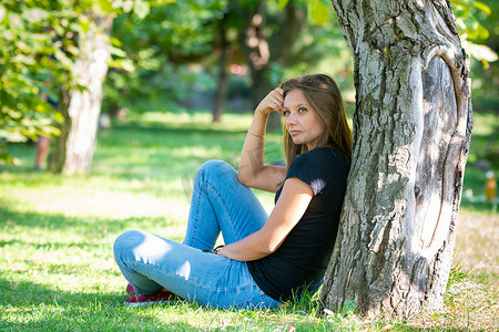 一个女孩坐在阳光明媚的公园的树下，望着远方沉思