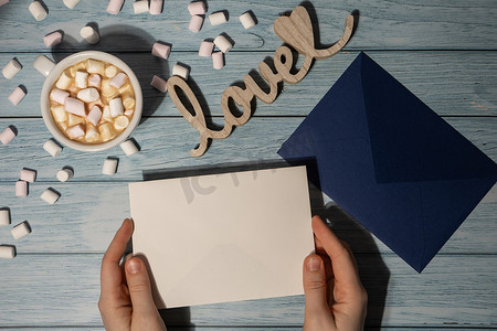 爱心手托卡通摄影照片_女手拿着空的问候语或邀请卡，用木制蓝色背景上的白色咖啡和棉花糖模拟蓝色信封。