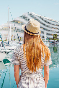 戴着帽子的金发女人背对着大海、游艇和小船背景站着。