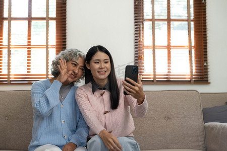 老人玩手机摄影照片_微笑的年长母亲和成年女儿一起使用电话，坐在家里舒适的沙发上，快乐的年轻女人和成熟的妈妈看着智能手机屏幕，看视频，玩得开心，两代人