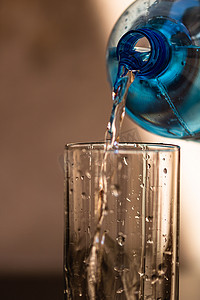 蓝色塑料瓶摄影照片_在模糊的背景下将蓝色塑料瓶中的水倒入玻璃杯中。
