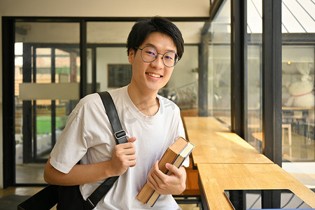 戴眼镜的亚裔男大学生拿着书，对着镜头微笑。