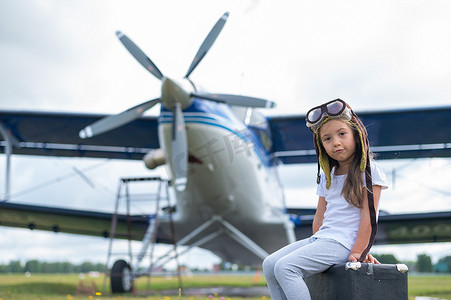 一个穿着飞行员服装的小女孩坐在一个手提箱上，背景是一架带螺旋桨的飞机。