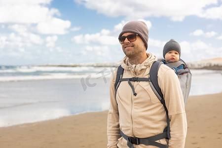 年轻的父亲在多风的沙滩上背着他的婴儿男婴儿子在背包里。