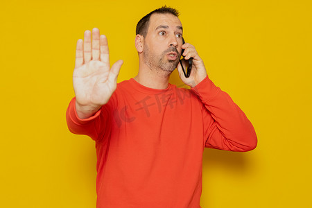 留着胡子的西班牙裔男子在手机上聊天时生气地用手在黄色背景下做了一个停车标志。