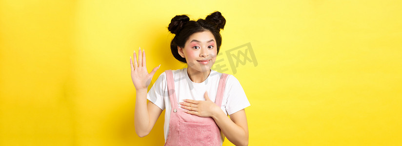 可爱的亚洲女孩的形象，化妆诚实，举手，放在心上，说实话，宣誓和微笑，站在黄色背景上
